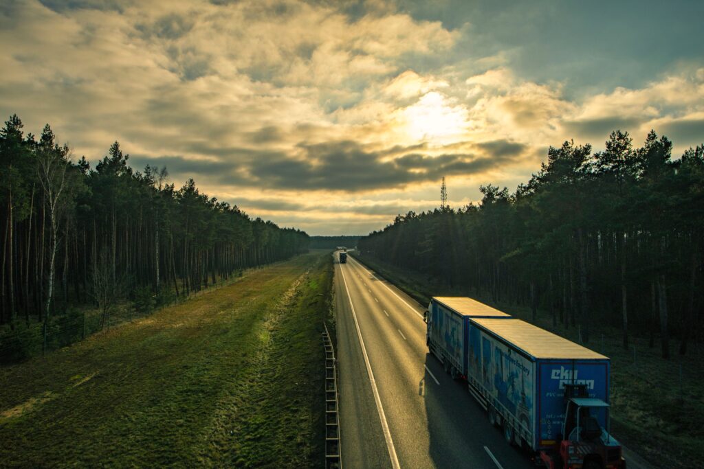 Trucks drive on a highway flanked by dense forest at sunset.
