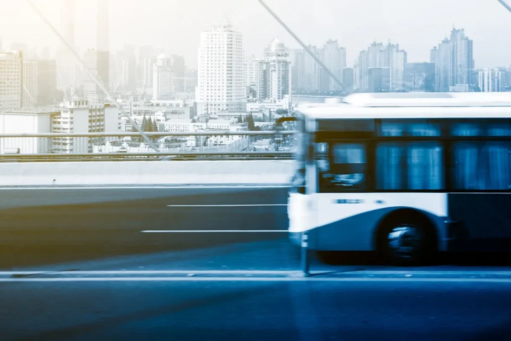 Imagem de um ônibus urbano em movimento sobre uma ponte, com prédios modernos ao fundo e uma cidade movimentada.