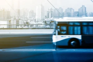 Imagem de um ônibus urbano em movimento sobre uma ponte, com prédios modernos ao fundo e uma cidade movimentada.