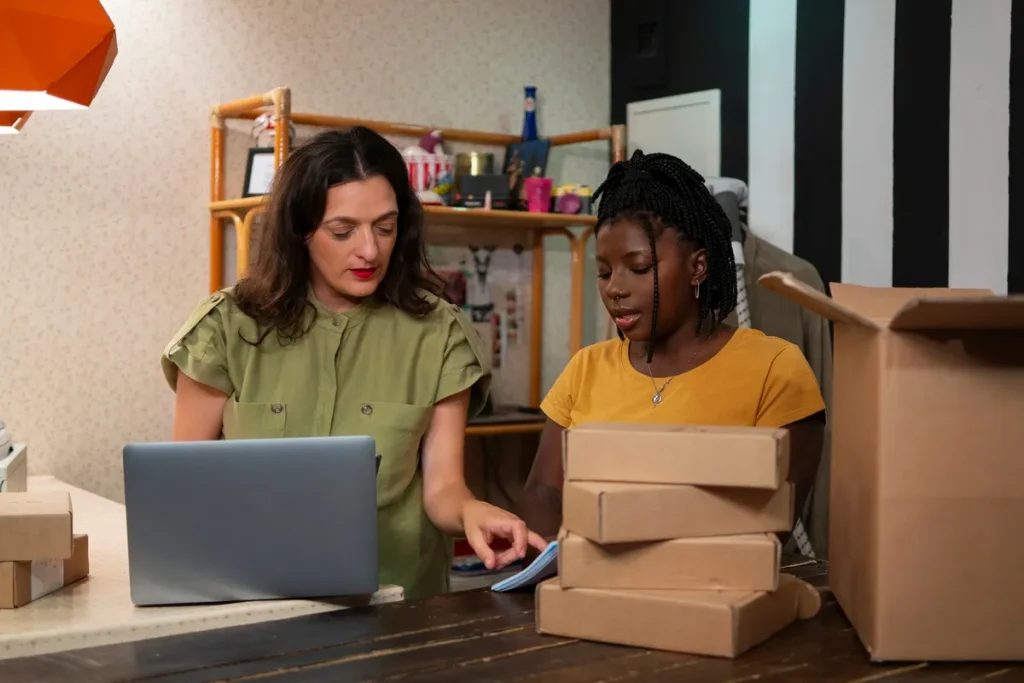 Duas mulheres organizando pacotes em ambiente de trabalho com notebook, representando logística e gestão de transporte de cargas pequenas.