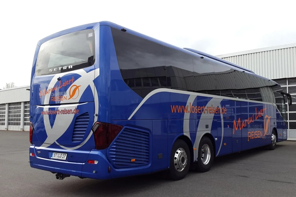 Ônibus de turismo azul da empresa Marcus Losert estacionado, com design moderno, três eixos e janelas escuras, em frente a um galpão