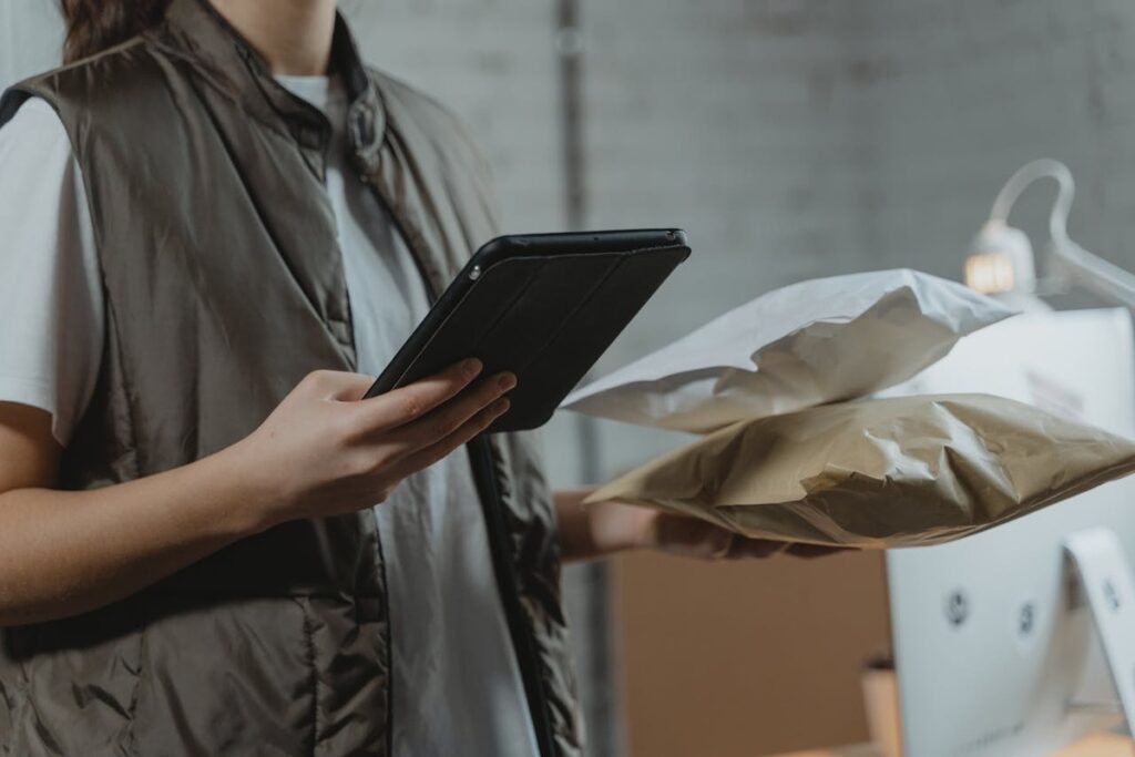Uma pessoa de colete segurando um tablet em uma mão e duas embalagens de papel ou plástico na outra, em um ambiente que sugere logística ou e-commerce, com foco na eficiência e escalabilidade de entregas.