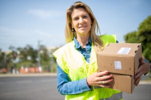 Uma mulher sorridente vestindo um colete de segurança verde limão e uma camisa jeans, segurando duas caixas de papelão, em um ambiente externo que sugere serviço de entrega ou logística.