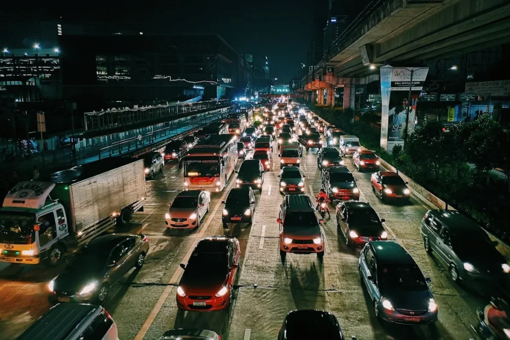 Vista noturna de uma movimentada avenida urbana com tráfego intenso e congestionado, iluminada pelas luzes dos veículos e dos edifícios ao redor, com um viaduto em destaque.