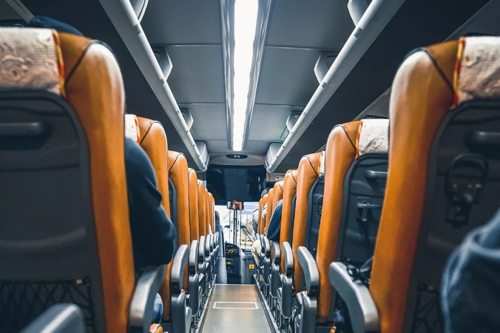 Interior de ônibus executivo com poltronas reclináveis em couro laranja e iluminação LED no teto, visto do corredor central