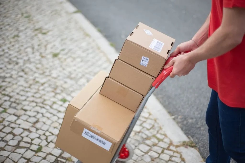 Um entregador em camisa vermelha empurrando um carrinho com várias caixas de papelão, simbolizando o serviço de remessa expressa e logística de entregas.