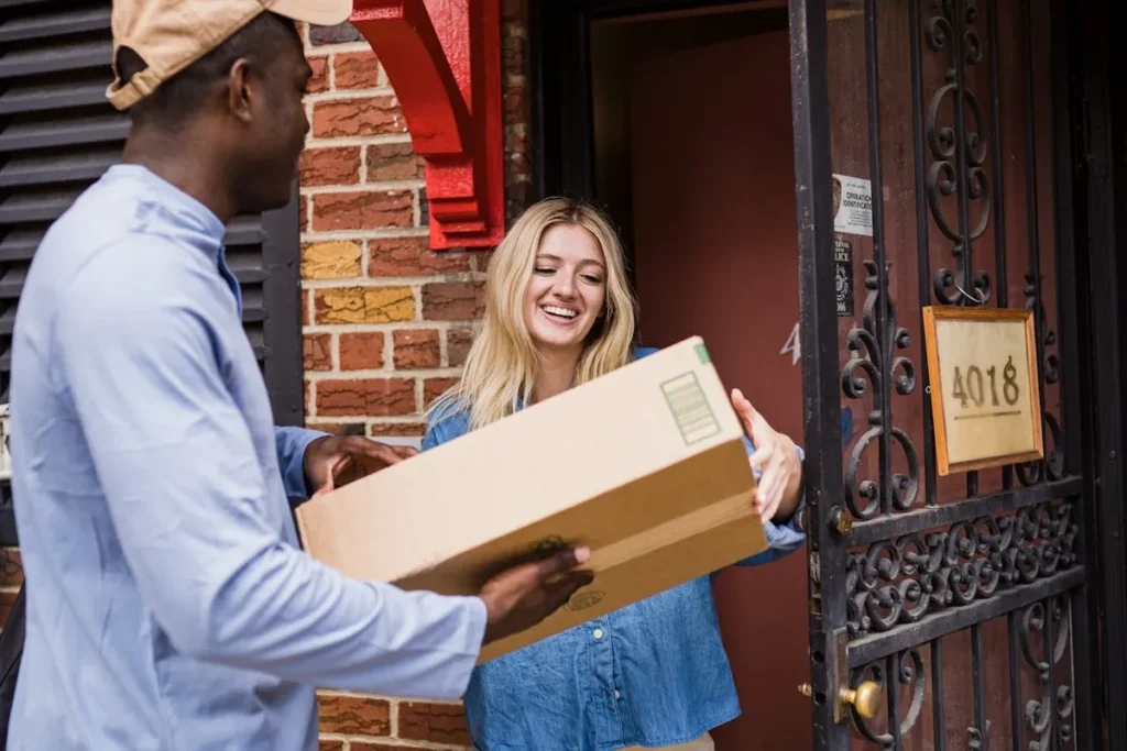 Um entregador sorridente entregando uma caixa de remessa expressa a uma mulher sorridente na porta de uma casa de tijolos, ilustrando a finalização de uma entrega rápida.