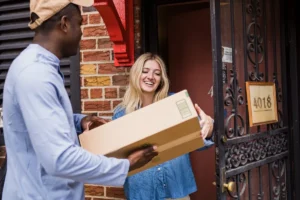 Um entregador sorridente entregando uma caixa de remessa expressa a uma mulher sorridente na porta de uma casa de tijolos, ilustrando a finalização de uma entrega rápida.