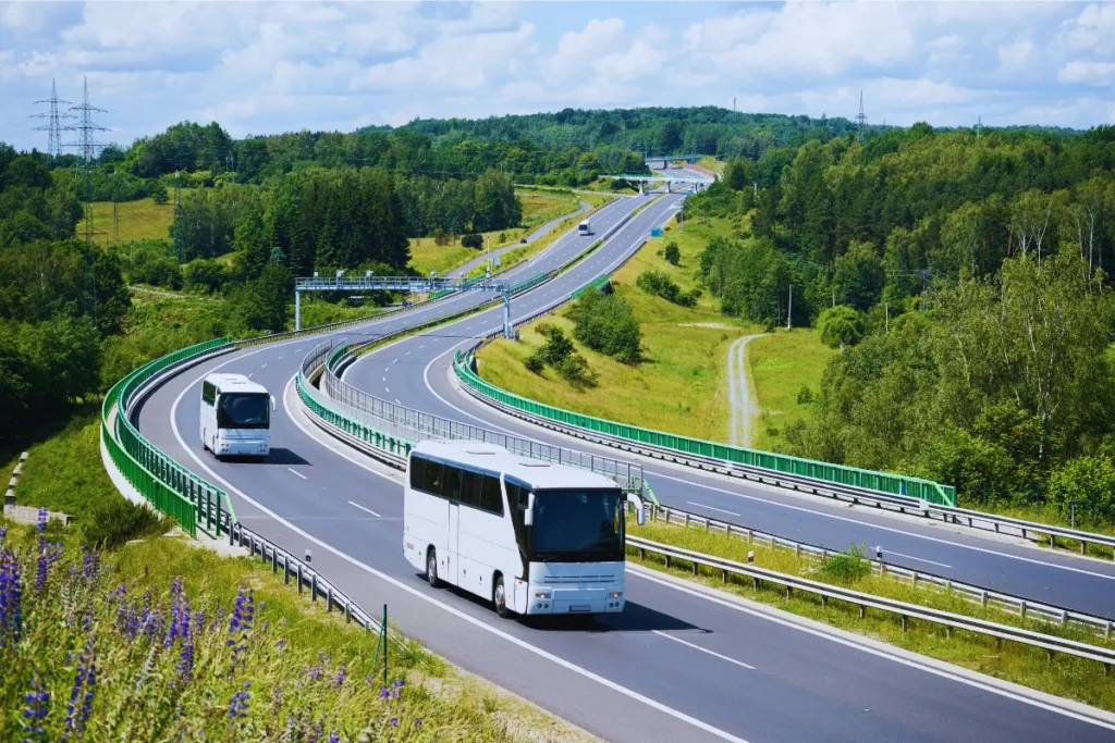 Dois ônibus brancos circulando por uma rodovia moderna com pistas largas e curvas suaves, cercada por vegetação e áreas verdes.