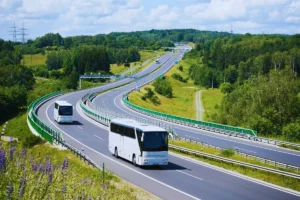 Dois ônibus brancos circulando por uma rodovia moderna com pistas largas e curvas suaves, cercada por vegetação e áreas verdes.