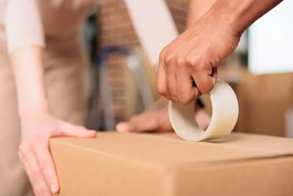 Duas pessoas fechando uma caixa de papelão com fita adesiva, mostrando o processo de acondicionamento de embalagens para transporte ou armazenamento.