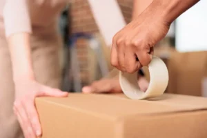 Duas pessoas fechando uma caixa de papelão com fita adesiva, mostrando o processo de acondicionamento de embalagens para transporte ou armazenamento.
