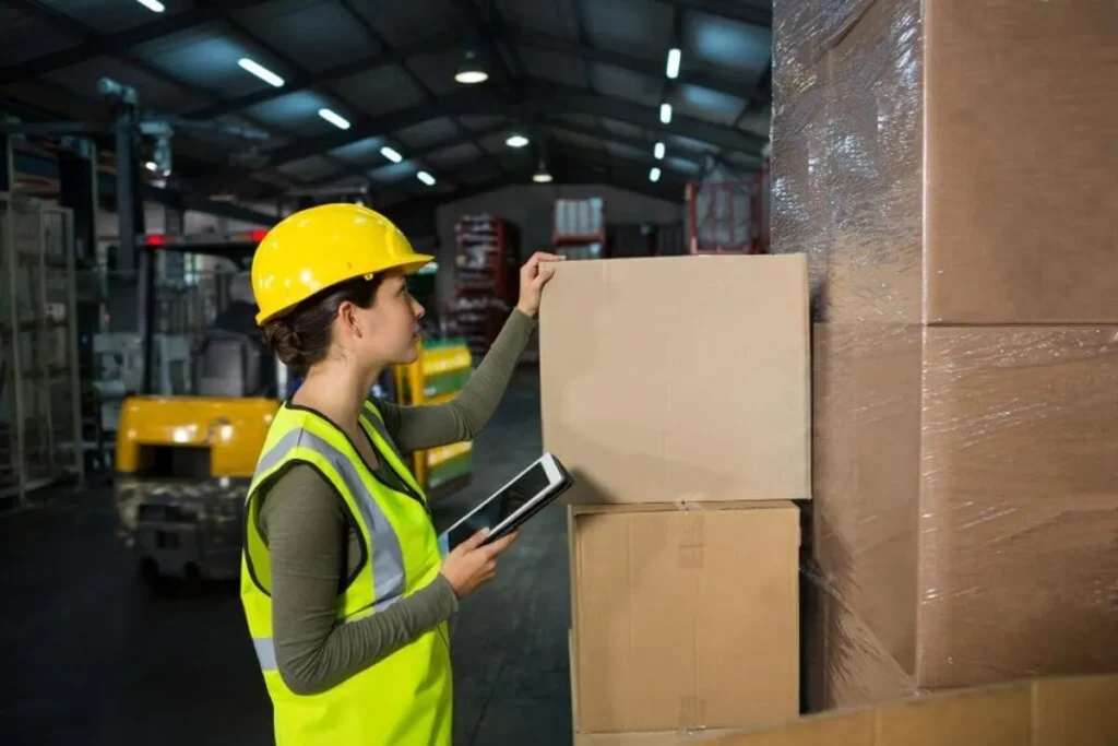 Mulher de capacete amarelo e colete de segurança, segurando um tablet em um galpão. A imagem representa o tema de logística, transporte e cubagem.
