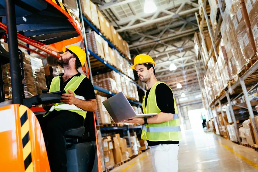 Dois trabalhadores de armazém, um na empilhadeira e outro com um laptop, verificando o inventário e a roteirização das operações.