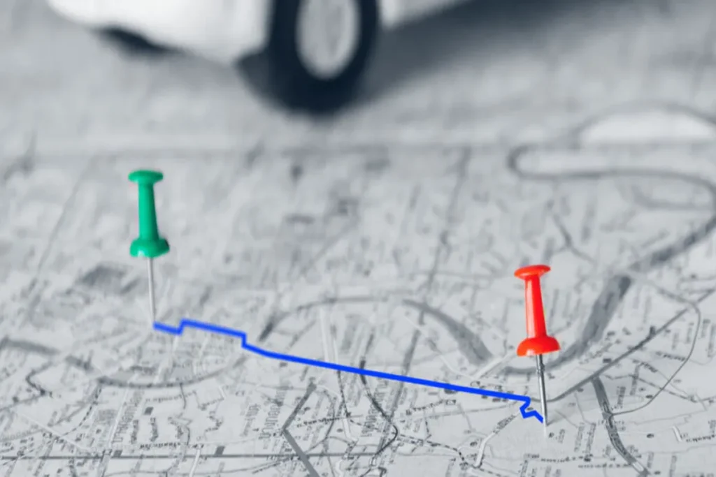 Um alfinete verde e um vermelho em um mapa com uma linha azul, representando a roteirização de um trajeto, com um carro de brinquedo desfocado ao fundo.