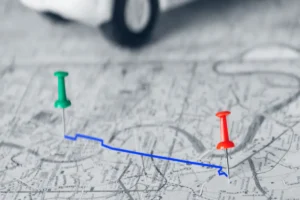 Um alfinete verde e um vermelho em um mapa com uma linha azul, representando a roteirização de um trajeto, com um carro de brinquedo desfocado ao fundo.