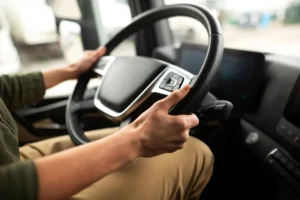Mãos de uma pessoa a segurar o volante de um camião. A cena está focada no volante e nas mãos, com a cabine e o exterior do veículo desfocados.