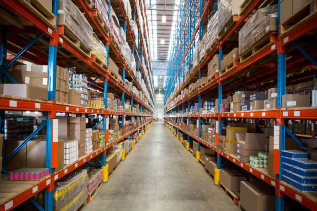 Vista interna de um grande armazém com longos corredores de prateleiras cheias de caixas e produtos, representando a logística de estoque.