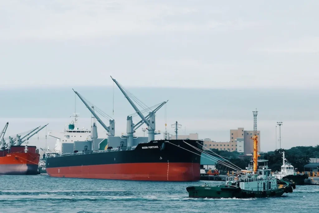 Vista de navio de carga geral 'NICOS FORTUNE' atracado em porto industrial, com guindastes a bordo e um rebocador em primeiro plano, representando o transporte marítimo global e frete FOB.