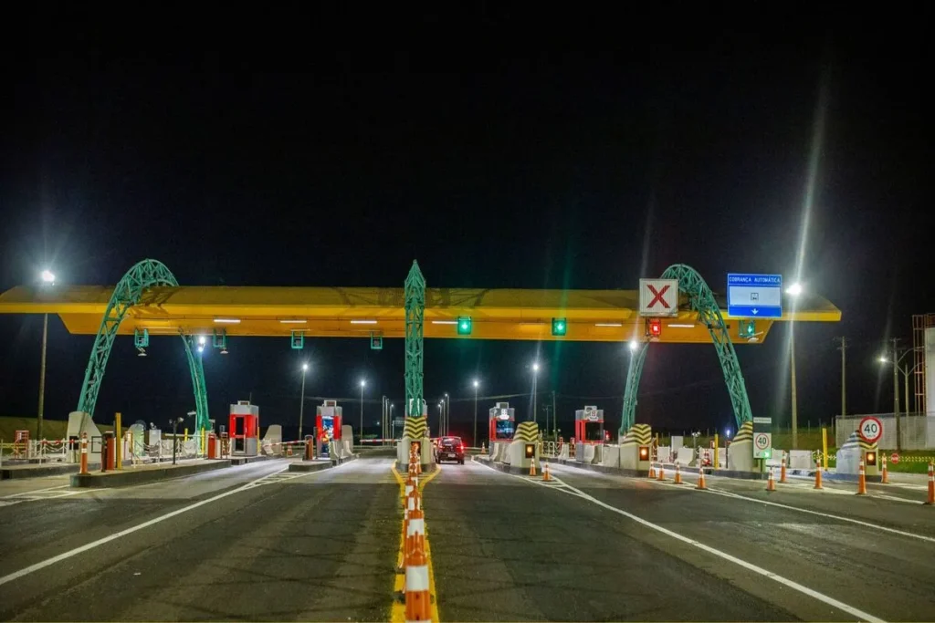 Uma praça de pedágio à noite, com um grande portal verde e amarelo sobre as faixas. As luzes de sinalização verde e vermelha estão ativadas, e um carro vermelho passa por uma das faixas.