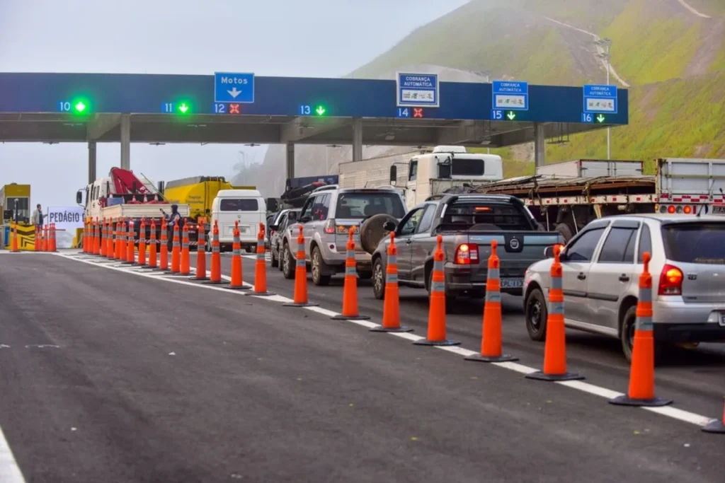 Carros, caminhonetes e caminhões em fila em uma praça de pedágio, com cones laranja delimitando as faixas. Placas de sinalização de cobrança automática e faixas com setas verdes e vermelhas estão visíveis, com montanhas enevoadas ao fundo.