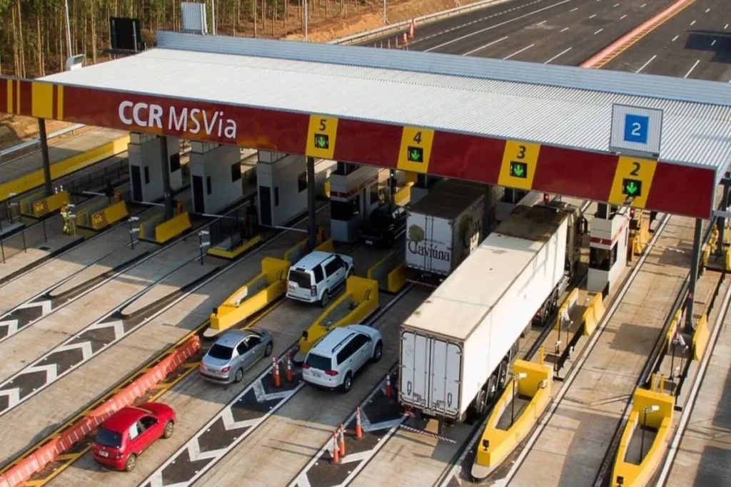 Vista aérea de uma praça de pedágio com o nome "CCR MSVia", mostrando várias faixas de tráfego com carros e caminhões, sinalizações com setas verdes e amarelas e barreiras de segurança.