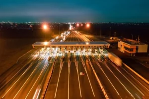 Vista aérea noturna de uma grande praça de pedágio, mostrando longas trilhas de luz de carros em uma rodovia de várias faixas. A estrutura do pedágio e suas cabines estão intensamente iluminadas.