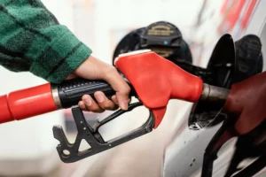 Mão segurando bico de abastecimento vermelho em posto de combustível para ilustrar preço do diesel.