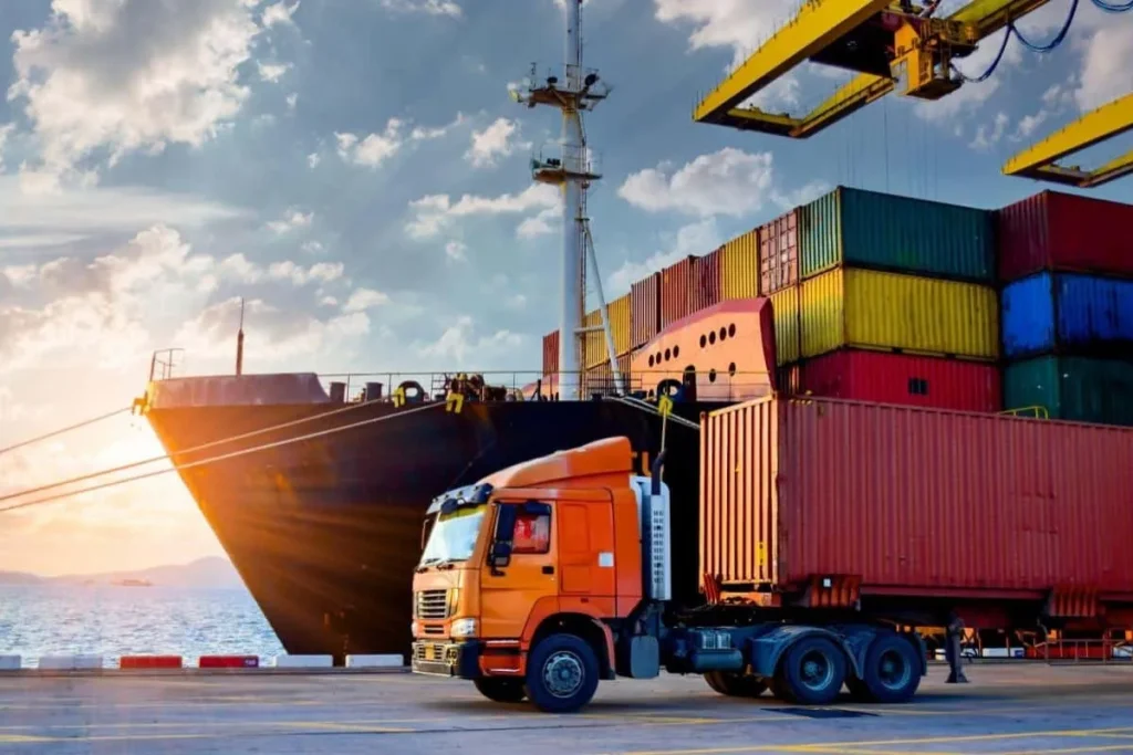 Navio cargueiro e caminhão transportando contêineres no porto, representando logística e supply chain internacional.
