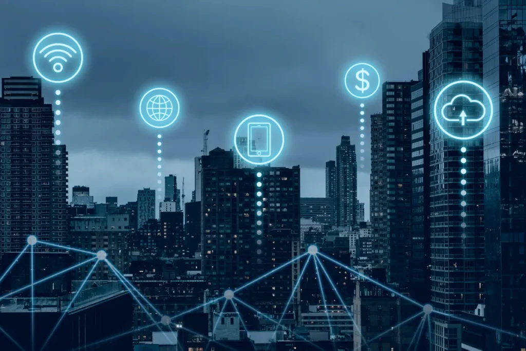 Vista em plano médio de edifícios altos de uma cidade em tons de azul escuro e ciano, com uma rede de pontos interconectados e linhas digitais brilhantes sobre a paisagem. Ícones luminosos, incluindo Wi-Fi, globo terrestre, smartphone/tablet, símbolo de dólar ($) e nuvem (cloud), flutuam sobre os prédios, representando a Internet das Coisas (IoT), comunicação global e transações financeiras digitais.