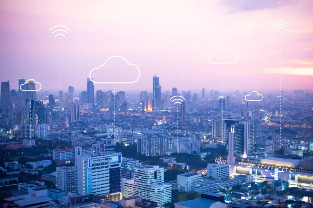 Vista aérea de uma grande metrópole ou paisagem urbana no crepúsculo, com luzes de edifícios altos. Sobre a imagem, ícones digitais em branco, como sinais de Wi-Fi e símbolos de nuvem (cloud), flutuam sobre os arranha-céus, representando a Internet das Coisas (IoT), a conectividade 5G e o armazenamento de dados em nuvem. A paleta de cores é um gradiente suave de roxo, rosa e azul.