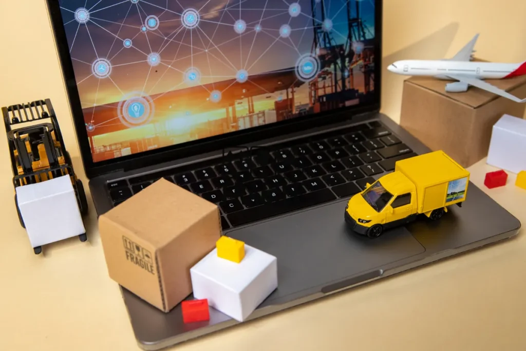 Composição digital mostrando o conceito de Internet das Coisas (IoT) aplicada à logística. Um laptop exibe uma tela com uma rede de pontos interconectados sobre um terminal de carga ao pôr do sol. Sobre o teclado, miniaturas de um caminhão de entrega amarelo, uma caixa de papelão e uma empilhadeira (forklift) representam a cadeia de suprimentos. Um modelo de avião está na lateral, simbolizando transporte aéreo.