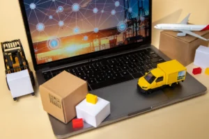 Composição digital mostrando o conceito de Internet das Coisas (IoT) aplicada à logística. Um laptop exibe uma tela com uma rede de pontos interconectados sobre um terminal de carga ao pôr do sol. Sobre o teclado, miniaturas de um caminhão de entrega amarelo, uma caixa de papelão e uma empilhadeira (forklift) representam a cadeia de suprimentos. Um modelo de avião está na lateral, simbolizando transporte aéreo.
