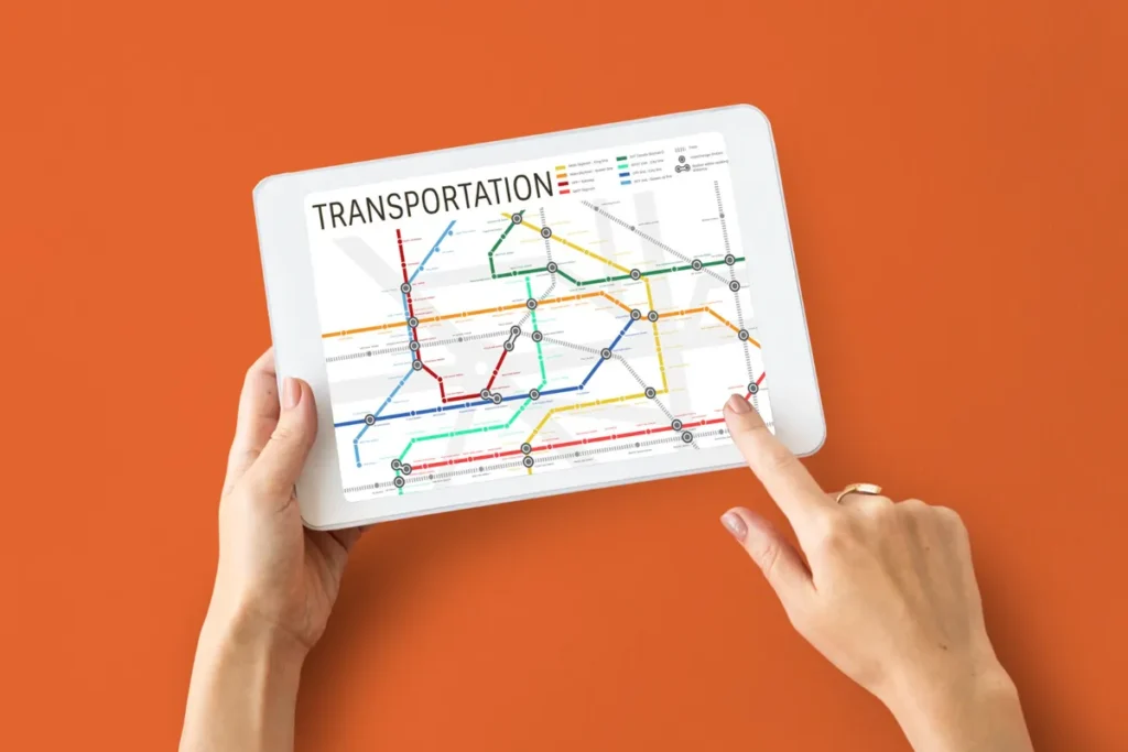 Duas mãos segurando um tablet branco contra um fundo laranja sólido. A tela do tablet exibe um mapa de transporte público ou logística de rotas com o título "TRANSPORTATION". O mapa é uma rede complexa de linhas coloridas (vermelho, azul, verde, amarelo) e pontos de conexão, simulando um sistema de rastreabilidade e planejamento de rotas. Um dedo está tocando a tela para interagir com o mapa.