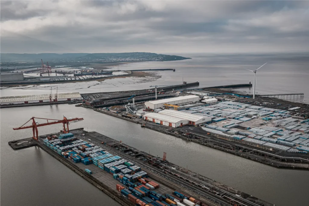 Vista aérea de um grande terminal portuário intermodal sob um céu nublado, mostrando guindastes, contêineres coloridos, pilhas de veículos brancos e turbinas eólicas na costa.