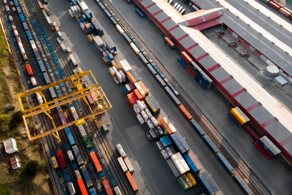 Vista aérea de um vasto terminal de transporte intermodal/ferroviário, repleto de contêineres de carga coloridos e guindastes de pórtico amarelos, ao lado de um grande galpão.
