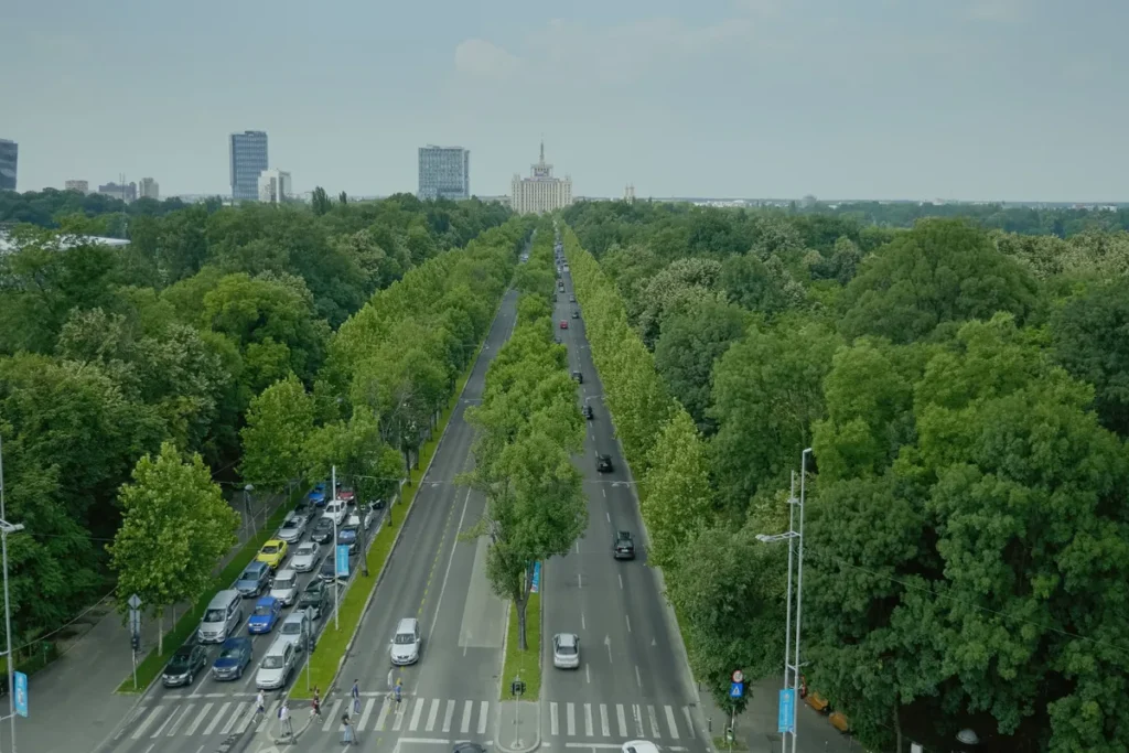 Vista aérea de uma grande avenida arborizada em uma cidade europeia (Bucareste), com tráfego leve e um grande edifício monumental ao fundo, destacando a integração de áreas verdes e infraestrutura urbana.